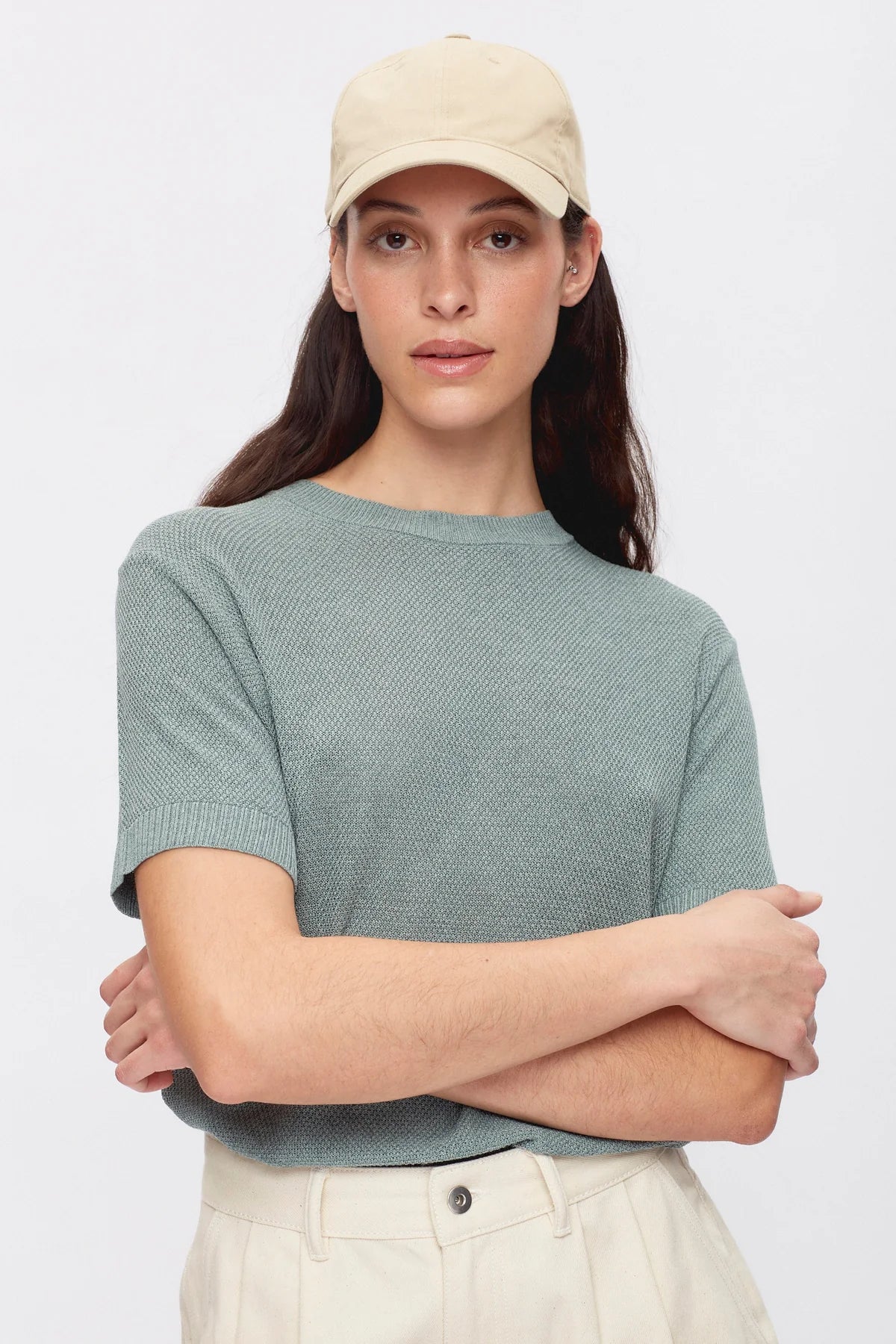 Woman wearing a light green cap and teal shirt with arms crossed on a white background